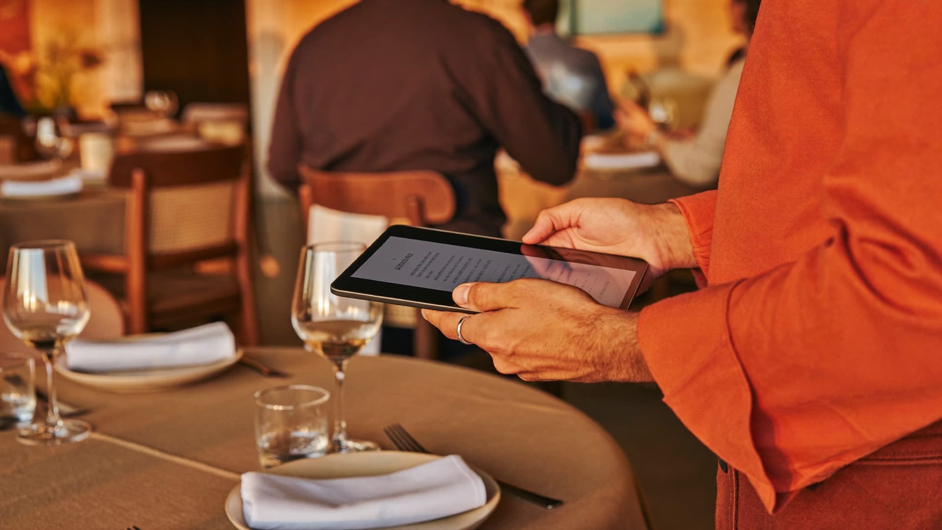 Las manos de un maître sostienen una tablet junto a una mesa puesta, con el movimiento de la sala desenfocado al fondo